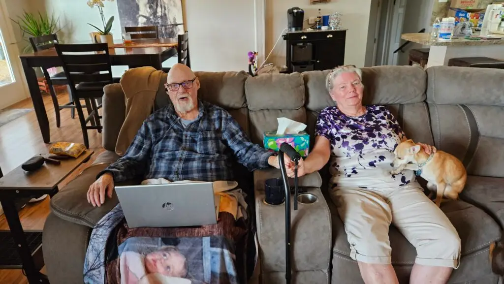 The last photo of my parents together, John (84) and Jackie Belville (79). They're sitting in a large plush brown sofa, Dad on the eft in a flannel shirt with a blanket over his legs and a computer on his lap, plus an oxygen tube for his nostrils. He's holding hands with Mom on the right, in a purple and white blouse with khaki capris. My blonde chihuahua Suzie is to Mom's left.