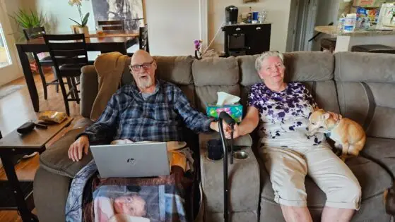 The last photo of my parents together, John (84) and Jackie Belville (79). They're sitting in a large plush brown sofa, Dad on the eft in a flannel shirt with a blanket over his legs and a computer on his lap, plus an oxygen tube for his nostrils. He's holding hands with Mom on the right, in a purple and white blouse with khaki capris. My blonde chihuahua Suzie is to Mom's left.