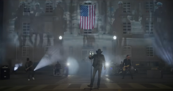Screenshot from Jason Aldean's "Try That in a Small Town" music video, showing the band staged at the Maury County, TN courthouse at night with lights projecting the image of four horseback riot cops.
